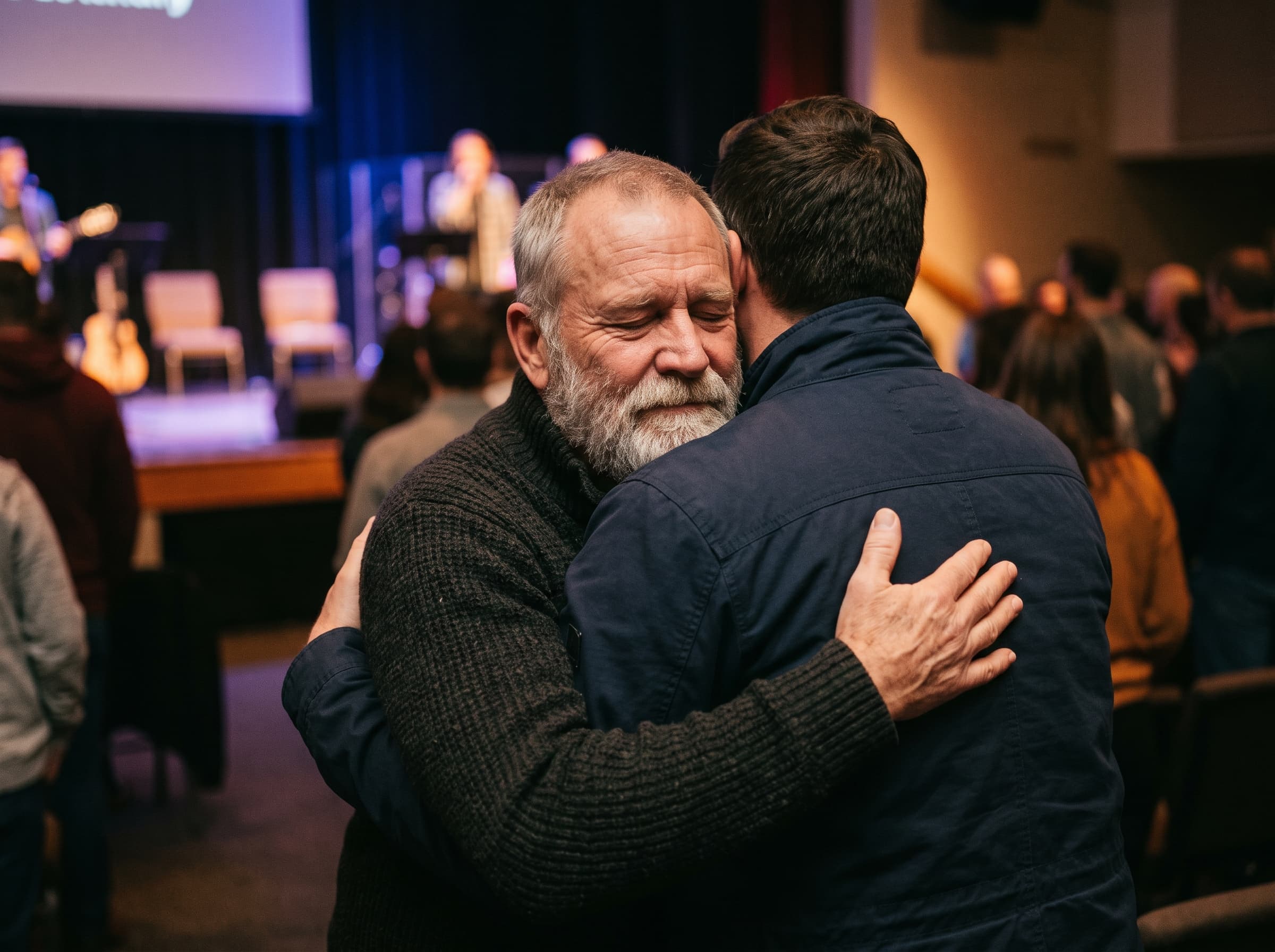 An older man with a grey beard closes his eyes while sharing a strong hug with a younger man.